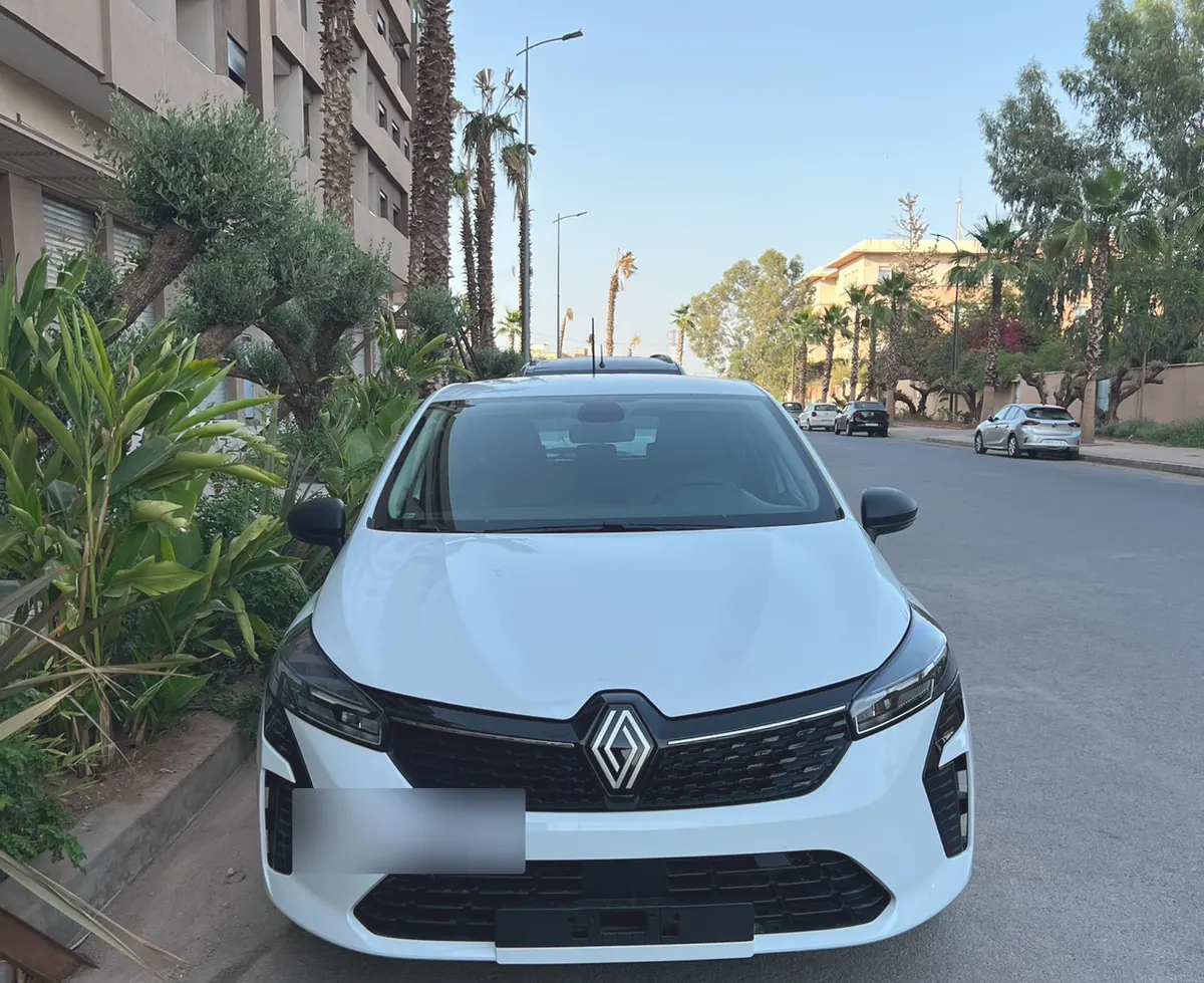 Renault Clio 5 blanche de l’agence, vue de face dans une rue bordée de palmiers, plaque floutée